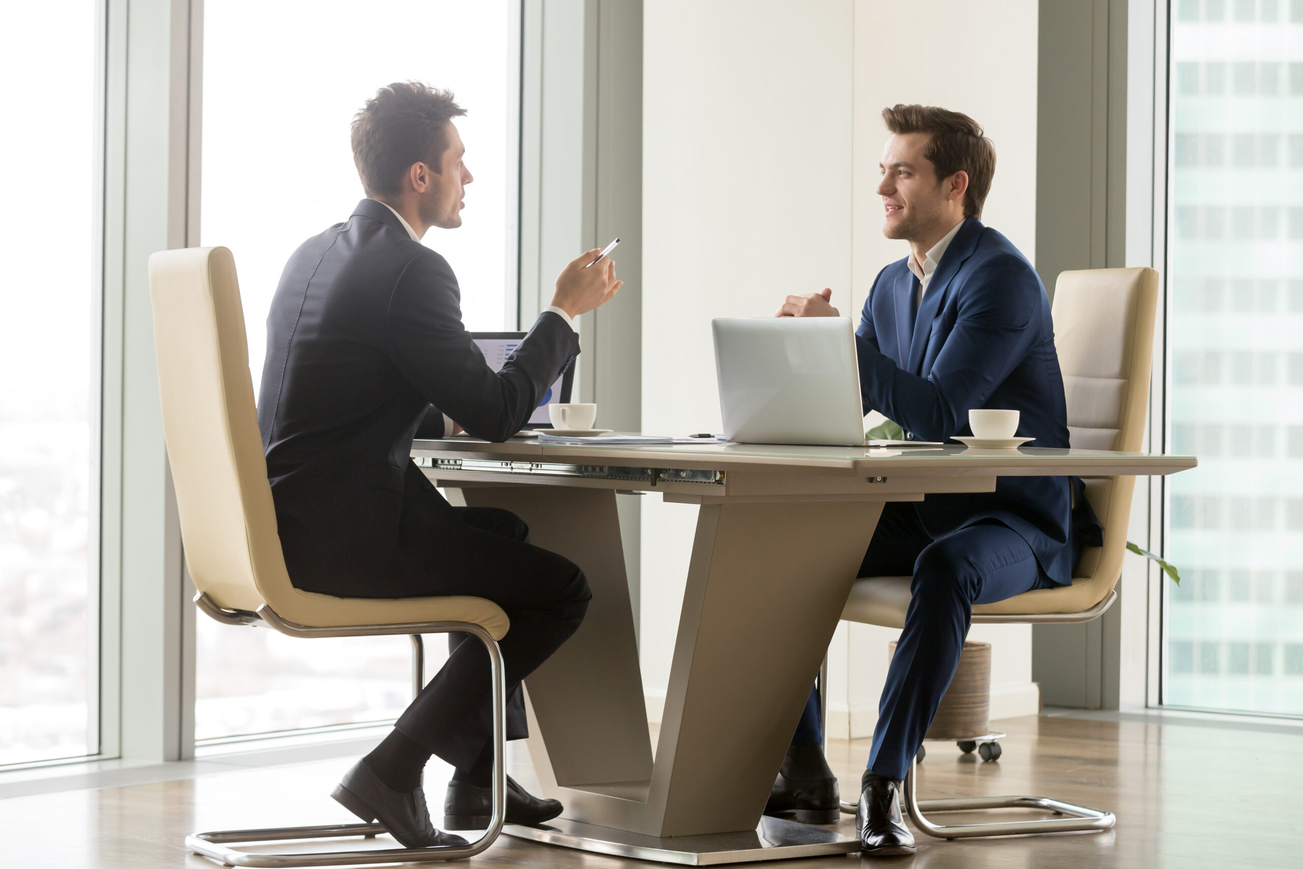 Successful entrepreneurs analyzing perspectives Two handsome businessmen sitting in comfortable chairs at desk with laptops in meeting room. CEO making important negotiation about companies partnership or corporate merger. Financiers planning deal